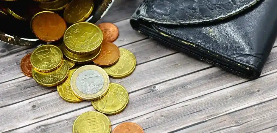 Un portefeuille en cuir usé et des pièces d'euro éparpillées sur une surface en bois.