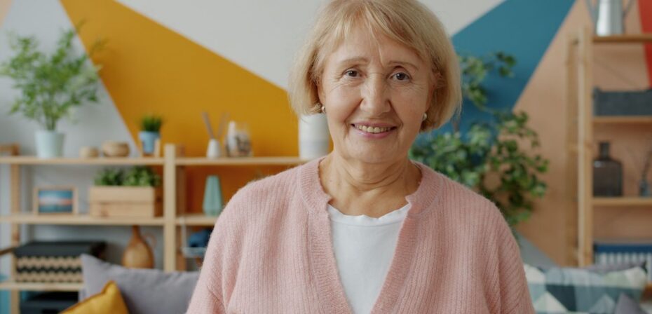 Une femme âgée sourit dans un salon chaleureux et coloré.